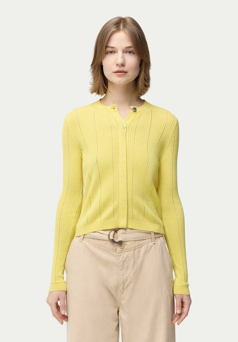 Gul ribbad kortärmad kofta med lång ärm, knäppning och en figursydd design. Bärs över beige byxor med en cirkulär bältesspänne i fokus.