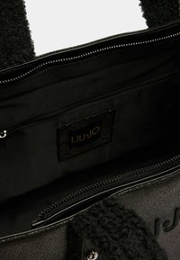 Sac fourre-tout noir en tissu texturé, avec fermeture éclair et doublure intérieure. Comprend une étiquette de marque et un accent doux et duveteux sur les poignées.