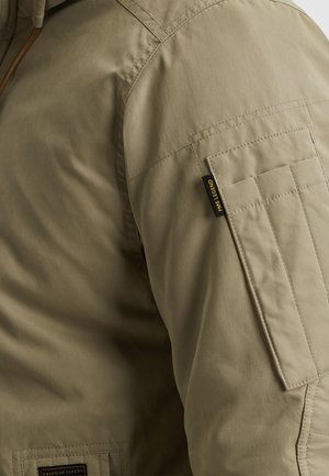 Olivovo zelená bunda s hladkou textúrou, ktorá má vrecko na ramene a detaily s vystuženým šitím. Na rukáve je viditeľná štítok s logom.