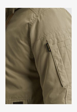 Giacca verde oliva con una texture liscia, dotata di una tasca sulla spalla e dettagli di cuciture rinforzate. Etichetta con logo visibile sulla manica.