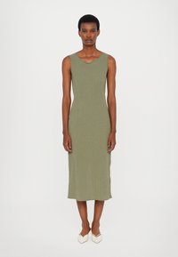 Femeie stând dreaptă, îmbrăcată într-o rochie midi fără mâneci, de culoare verde măsliniu, cu dungi, și pantofi albi cu vârful ascuțit, pe un fundal deschis și simplu.