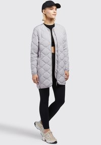 Veste matelassée gris clair avec un motif de losanges, fermeture éclair devant et poches latérales, associée à des leggings noirs et des baskets beiges.