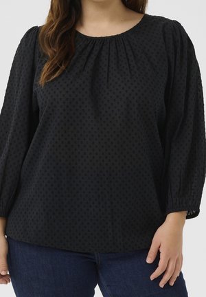 Vrouw die een zwarte, gestructureerde blouse met lange mouwen en een geplooide halslijn draagt, samen met donkerblauwe spijkerbroek, tegen een effen achtergrond.