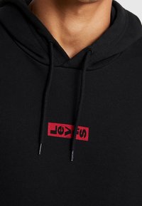 Sweat à capuche noir avec un logo rectangulaire rouge Levi's. Il est doté d'une capuche avec cordon de serrage et de poignets côtelés pour un ajustement confortable.