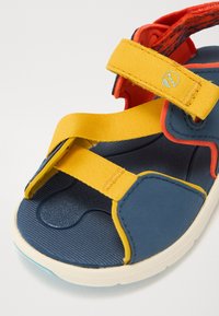 Sininen ja keltainen sandaali, jossa on teksturoitu kumipohja, kankaiset hihnat ja pehmustettu kantapää. Varusteena säädettävät keltaiset Velcro-hihnat ja logoyksityiskohta.
