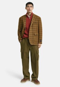 Bruin geruite blazer, rode kabelgebreide trui, olijfgroene cargobroek, bruine schoenen met dikke zolen. Heeft zakken op de broek en contrasterende texturen.