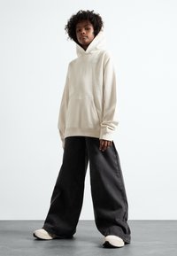 Beige kapucnis pulóver, elülső zsebbel, óriási fekete farmernadrággal és krém színű sportcipővel párosítva, semleges háttér előtt.