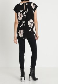 Blusa negra con estampado floral y mangas cortas, con un corte en la espalda tipo ojo de cerradura, de tela suave, y un estampado floral en blanco y rosa. Combinada con jeans ajustados negros y botas de tacón.