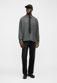 Άνδρας που φοράει φαρδιά γκρι πουκάμισο με κουμπιά, μαύρη γραβάτα, μαύρο παντελόνι, γυαλιστερά μαύρα παπούτσια και μαύρο καπέλο μπέιζμπολ, στέκεται μπροστά σε ένα απλό φόντο.