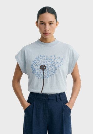 Mujer con cabello oscuro, con una camiseta sin mangas azul clara con un estampado de diente de león azul, y pantalones azul marino de talle alto con las manos en los bolsillos.
