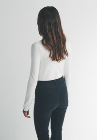 Top alb cu mâneci lungi, asortat cu blugi închiși la culoare, cu talie înaltă, evidențiind o siluetă ajustată și o textură netedă. Părul cade până sub umeri.