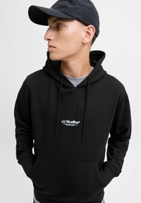 Schwarzer Kapuzenpullover mit einer Vorderasche, Kordeln und weißem, gesticktem "JJ Studios"-Logo. Der Stoff wirkt weich und matt.