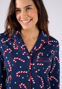 Camisa de pijama azul marino con estampado de bastones de caramelo y corazones. Cuenta con ribete rojo, cuello con muesca y un bolsillo en el pecho con cierre de botón.