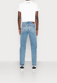 Vaqueros de mezclilla azul claro con corte recto, con dos bolsillos traseros y una marca de cuero marrón en la pretina. Combinados con una camiseta blanca.