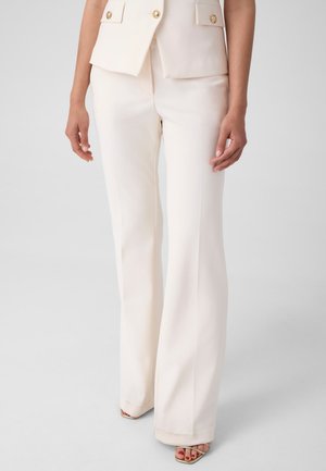 Woman wearing high-waisted, wide-leg cream pants paired with a matching cream vest and beige open-toe heels.