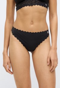 Juodas bikinis su banguotais kraštais, pagamintas iš švelnios medžiagos; pasižymi glaudžiu prigludimu ir mažu logotipo elementu šone.