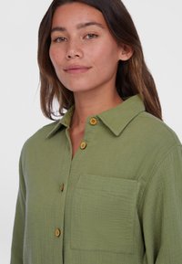 Grüne Hemdjacke aus strukturiertem Stoff mit spitzem Kragen, einer Brusttasche und Holzknöpfen. Eng anliegend am Hals.