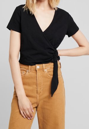 Frau, die ein schwarzes Wickeloberteil mit kurzen Ärmeln und seitlicher Schnürung sowie hoch geschnittene beige Hose trägt und mit einer Hand in der Hüfte posiert.