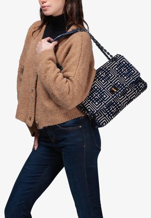 Borsa in tessuto intrecciato con un motivo geometrico blu navy e crema, dotata di una chiusura in tonalità oro e di una tracolla a catena. Abbinata a un cardigan color cammello e a jeans scuri.