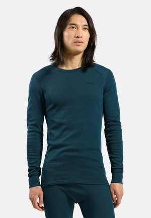 Man with long dark hair wearing a fitted dark teal long-sleeve shirt and matching pants, standing against a plain background.