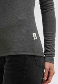Gray long-sleeve top made of soft fabric, featuring a slim fit, ribbed texture, and a white "SENSES" label on the side. Paired with black jeans.