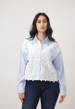 Donna con capelli lunghi e scuri che indossa una camicia a righe blu sovrapposta a un top bianco in pizzo floreale con bottoni, abbinata a jeans blu scuro.