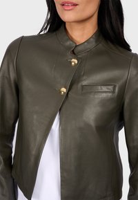 Olivově zelená kožená bunda s vysokým límcem, asymetrickým předním dílem, dvěma zlatými knoflíky a malou kapsou na prsou. Hladký povrch.