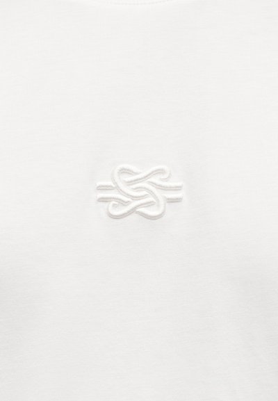 Λευκό βαμβακερό μπλουζάκι με διακριτικό λευκό κεντητό σχέδιο κόμπου στο στήθος, που παρουσιάζει ομαλή υφή και μινιμαλιστικό στιλ.