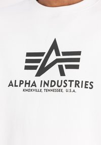 Bijela dukserica s crnim logotipom sa stiliziranim slovom "A" i horizontalnim prugama. Natpis glasi "ALPHA INDUSTRIES, KNOXVILLE, TENNESSEE, SAD."