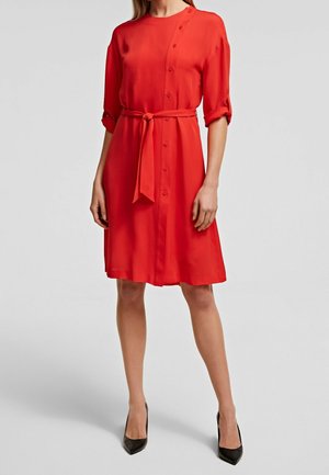 Rode jurk met een ceintuur in de taille, net boven de knie, met knoopdetails en opgerolde mouwen. De stof lijkt glad en lichtgewicht.