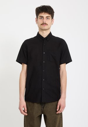 Junger Mann mit kurzen braunen Haaren und Schnurrbart, der ein schwarzes, kurzärmeliges Hemd mit Knöpfen und braune Hosen trägt, vor einem schlichten Hintergrund.