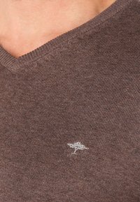 Brown knit sweater with a V-neck design, featuring a subtle silver tree logo on the chest and textured detailing along the neckline.