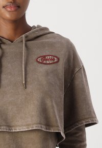 Hnědá zkrácená mikina s texturovanou tkaninou a vyšitým logem "Guess" v červené barvě na světle hnědém oválném štítku. Součástí je kapuce se šňůrkami.