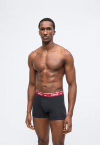 Homme musclé debout, portant des boxers Nike noirs avec une ceinture rouge, expression neutre, fond gris clair uni.