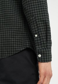 Dlouhý rukáv košile s tmavě zeleno-černým kostkovaným vzorem, s bílými knoflíčky na zápěstí, nad černými kalhotami a viditelnou rukou.