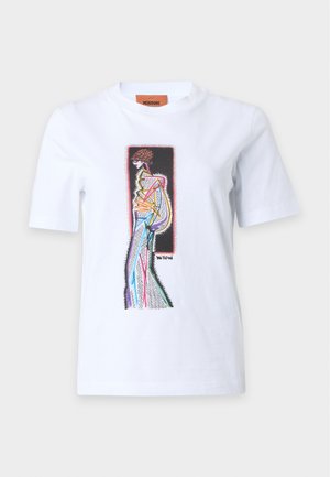 Frau mit langen dunklen Haaren, die ein weißes Grafik-T-Shirt und schwarze Zickzack-Hosen trägt, hält eine große schwarz-weiß gemusterte Tasche.