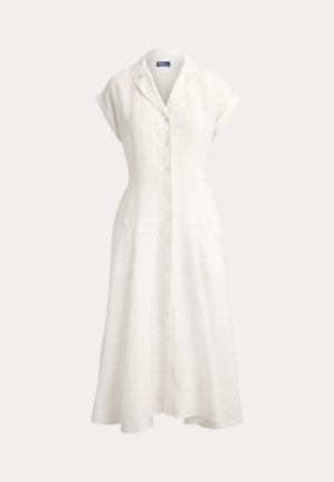 Mujer con el pelo trenzado, vestida con un vestido midi blanco con botones, mangas cortas y sandalias negras planas, de pie con las manos en los bolsillos.