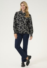 Blusa nera con motivo a catena, maniche lunghe e colletto, abbinata a jeans scuri e calzature nere.