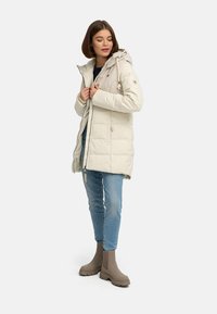 Woman wearing a beige hooded puffer coat, blue jeans, and taupe ankle boots standing against a plain white background.