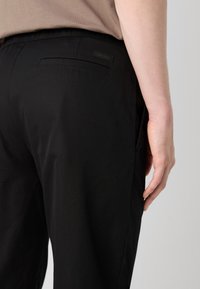 Sorte bukser med en glat tekstur, som har sidelommer og et strømlinet design. En lille Calvin Klein-label er synlig på bagsiden.