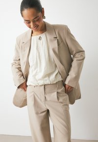 Blazer beige ajustado con pantalones a juego, combinado con una blusa blanca texturizada. Presenta un corte relajado y detalles sutiles en los bolsillos.