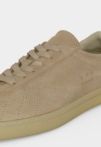 Baskets en daim beige avec lacets ton sur ton, motifs géométriques cousus, col rembourré et semelle en caoutchouc, vue partielle latérale.