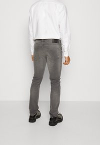 Man som bär åtsittande grå jeans, svarta skor och en vit långärmad skjorta, står mot en enkel ljus bakgrund med ryggen vänd.