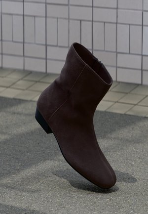 Bottine en daim marron foncé à talon bas, positionnée debout sur un pavé texturé contre un mur carrelé.