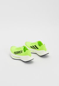Neon zöld sportcipők fekete kiegészítőkkel, hálós felső résszel, párnázott talppal és merész "TERREX" márkázással az oldalon.