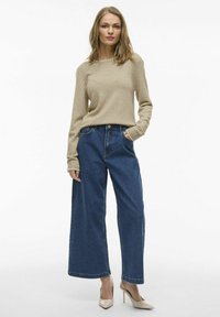 Suéter beige de manga larga, corte holgado, con puños acanalados. Pantalones de mezclilla azul de pierna ancha con bolsillos frontales, combinados con zapatos de tacón beige.