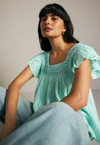 Lyse turkise bluse med firkantet udskæring, der har et detaljeret hækledesign og flæset korte ærmer. Blødt, struktureret stof.