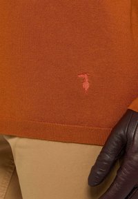 Oranger gestrickter Pullover mit geripptem Saum und kleinem roten Logodetail; getragen mit beigen Hosen und einem dunklen Lederhandschuh an der rechten Hand.