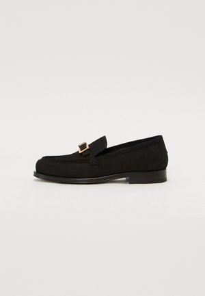 Black suede loafer with a low heel, featuring a gold-tone square buckle accent and stitching details along the upper. Rounded toe design.