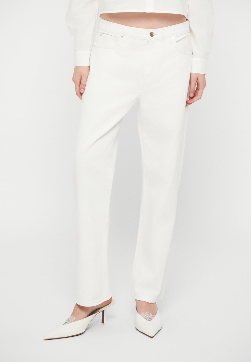 Jean en denim blanc avec une coupe droite, présentant des poches avant et une fermeture à bouton métallique, associés à des talons aiguilles blancs.